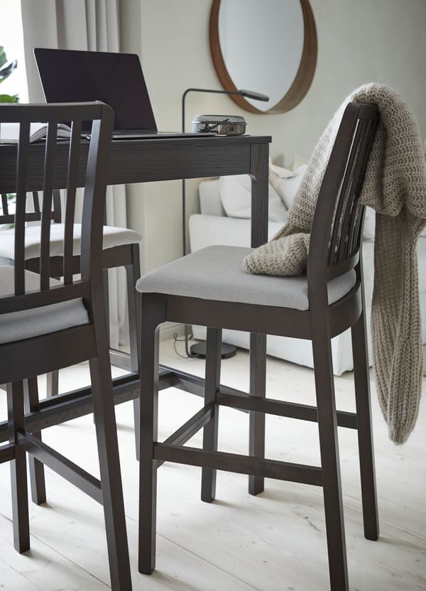 An IKEA EKEDALEN dark brown bar table and bar chairs with light grey cushions, with an open computer on the table.