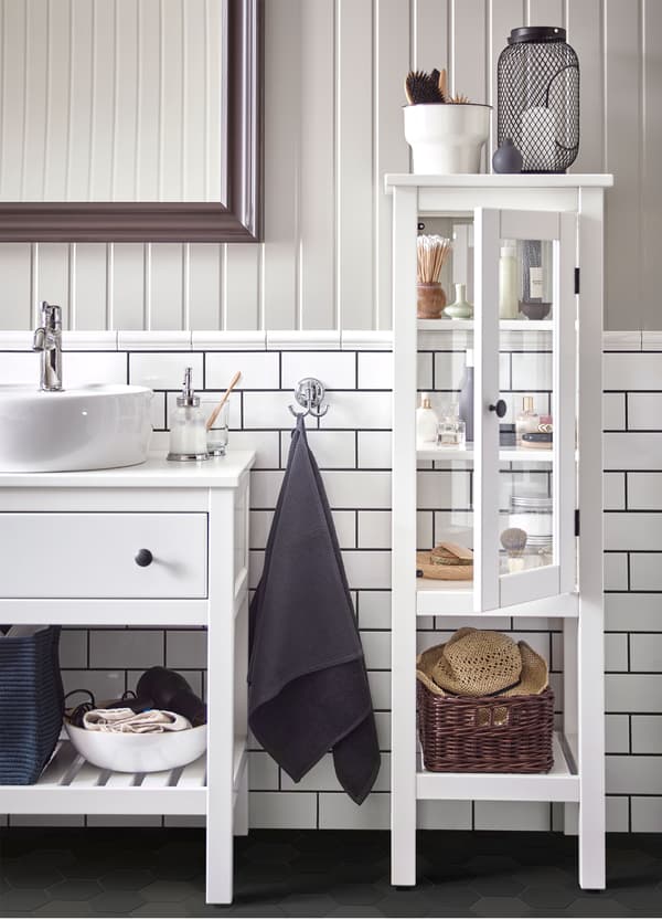 An IKEA HEMNES white high cabinet with glass doors, filled with bathroom accessories, beside a wash-stand.