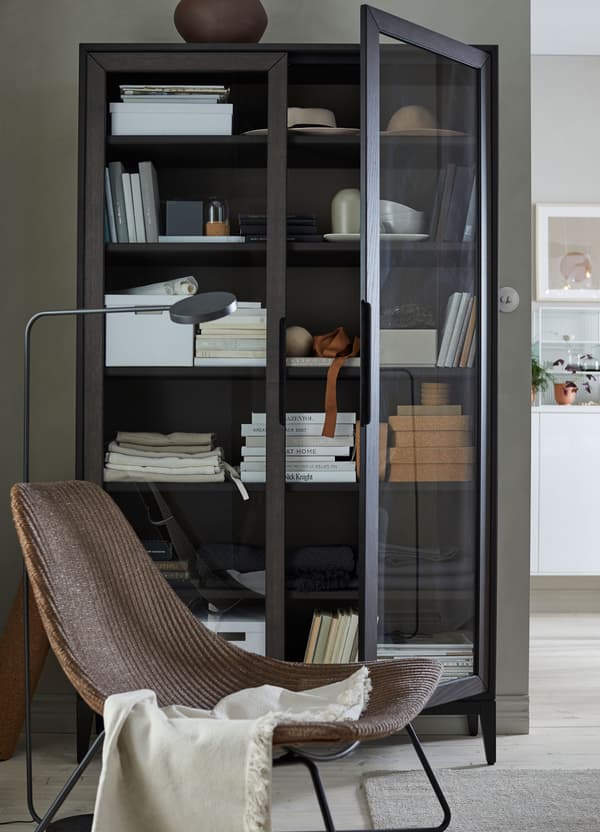 An IKEA REGISSÖR black brown glass door cabinet filled with boxes, books and glassware, behind a chair.