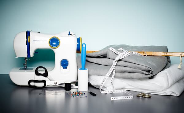 An IKEA sewing machine, fabric, measuring tape, pins, scissors and cotton thread on a table close to a wall.