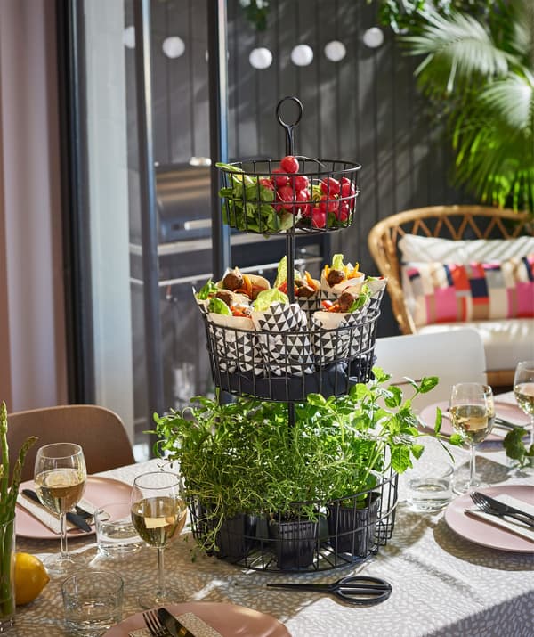 An IKEA SOMMAR 2018 3-tier serving basket in black steel on a dining table, filled with herbs and vegetables.