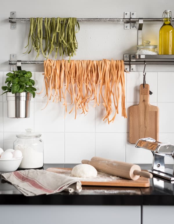 An IKEA wall mounted kitchen rail that is hanging green and orange coloured linguine to dry above a kitchen countertop.