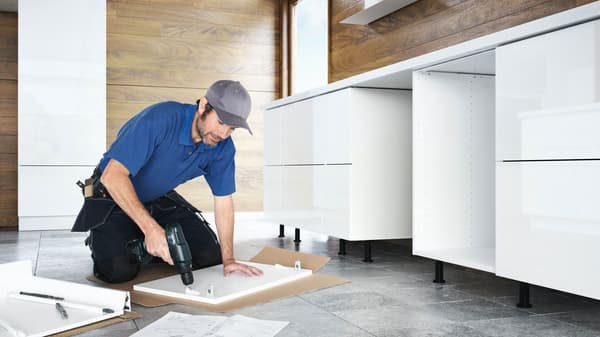 An image of a man in blue shirts assembling kitchen furniture using a power tool.