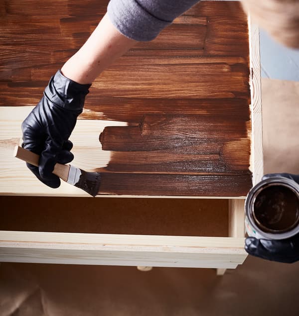 An interior designer wears black latex gloves while staining an IKEA RAST 3 drawer chest in dark brown.