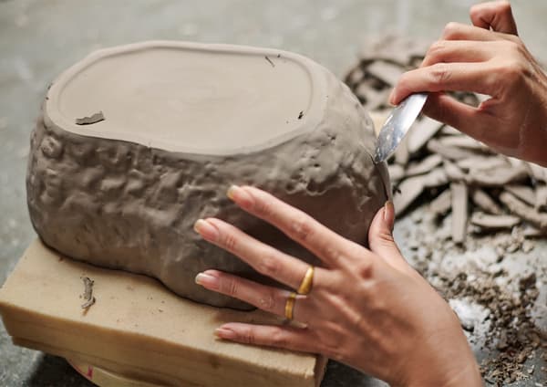 An irregular ceramic bowl in brown clay being perfected with a knife by hand by an artisan.