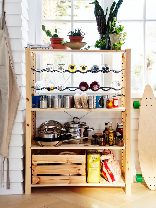 An IVAR unit stands by a window, organised as a small pantry with pots, dry goods, canned food, and bottles on racks.