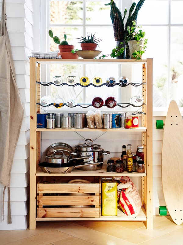 An IVAR unit stands by a window, organised as a small pantry with pots, dry goods, canned food, and bottles on racks.