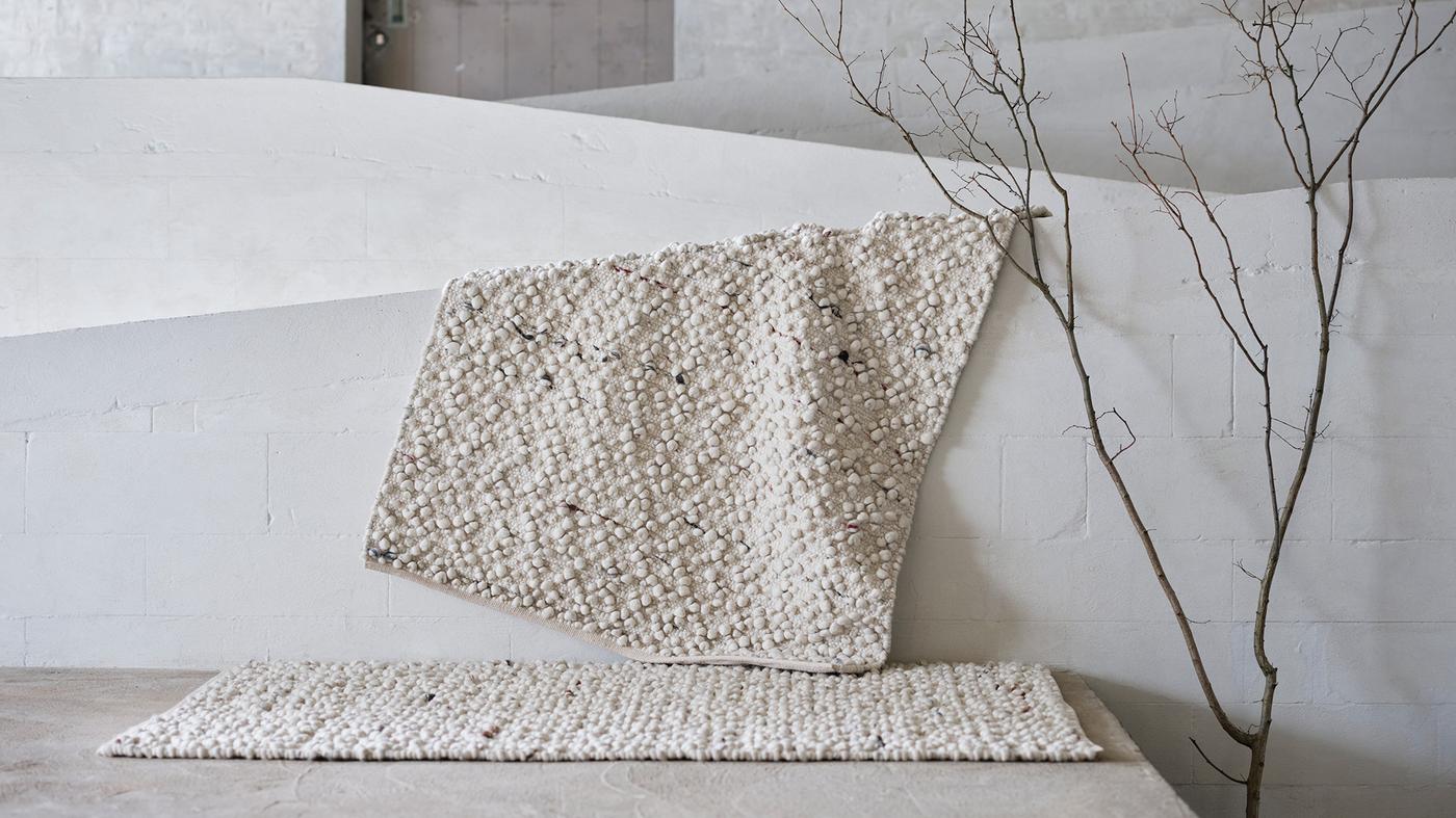 An off-white SVÄRDBORG rug with multicoloured yarns is draped over a white wall; another SVÄRDBORG rug lies in front.