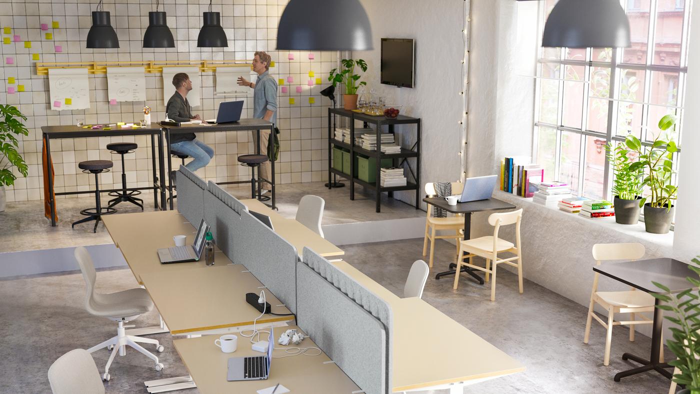 An open office with two men working by high tables, and a woman sitting on a beige/white chair by a desk writing emails.