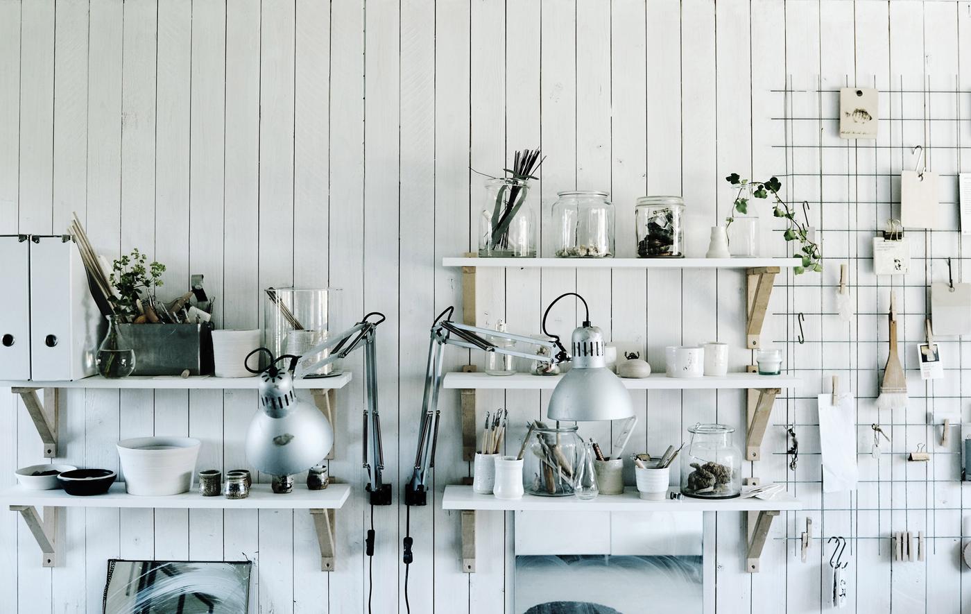 An open pantry with white shelves that hold clear jars and glassware and a wire rack with clip-on utensils and papers.