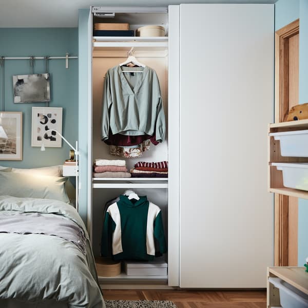 An open PAX wardrobe with white sliding doors has a white stained oak effect interior with clothes, baskets and drawers.