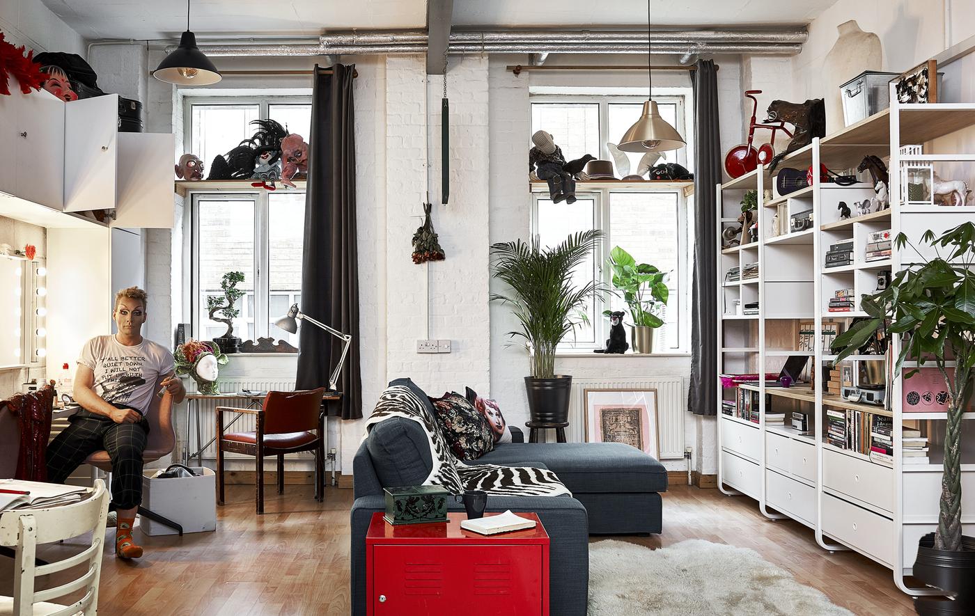 An open-plan living area with high ceilings, L-shaped sofa, wall storage unit and Donald sat on chair in front of mirror.