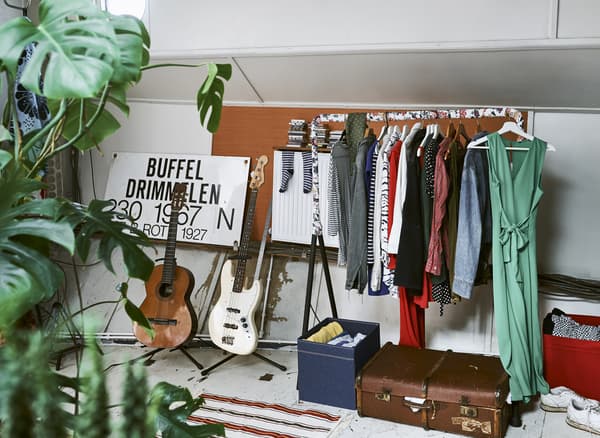 An open rail filled with clothes, next to two guitars on stands, a suitcase and some storage boxes.