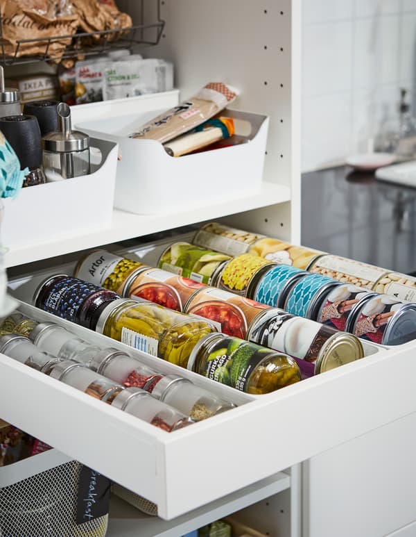 An open white drawer and shelves in a kitchen cabinet with neatly organized cans, spices and jars.
