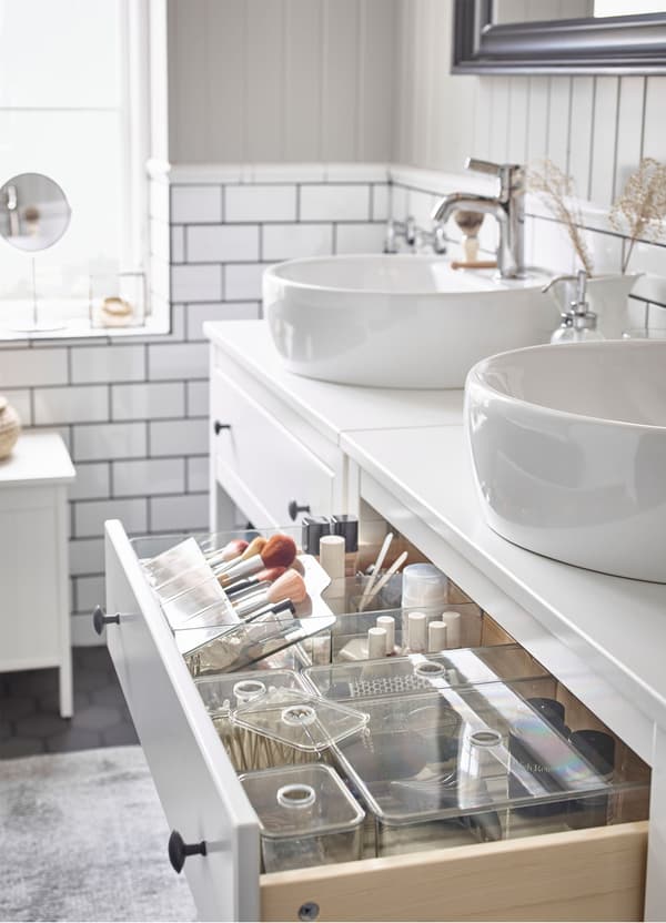 An opened drawer of a white wash-stand, revealing IKEA GODMORGON clear boxes with lids, filled with bathroom accessories.