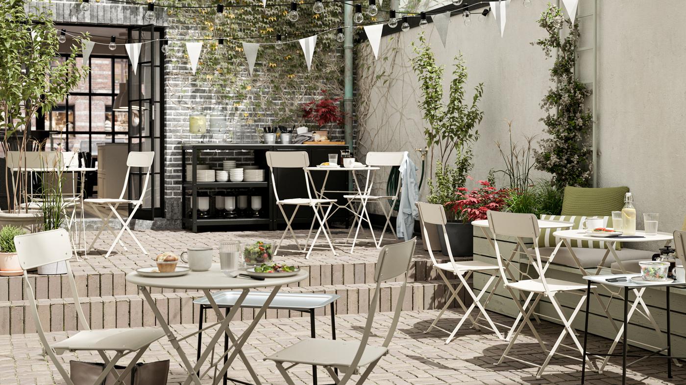 An outdoor cafe with beige steel foldable tables and chairs, white bunting, tiled floors and a serving station.