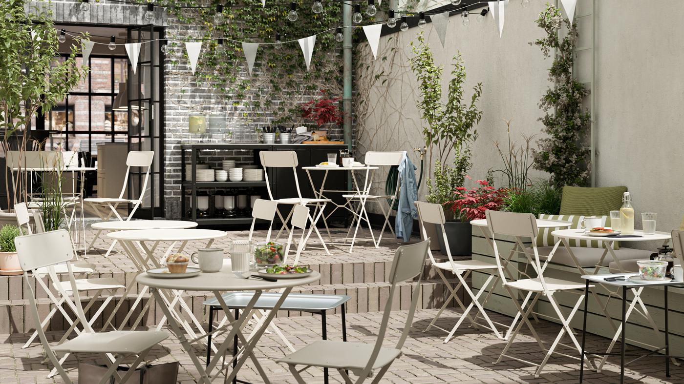 An outdoor cafe with beige steel foldable tables and chairs, white bunting, tiled floors and a serving station.