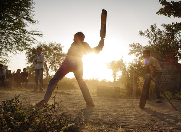 An outdoor evening scene in the countryside with several teenagers playing a baseball-like game.