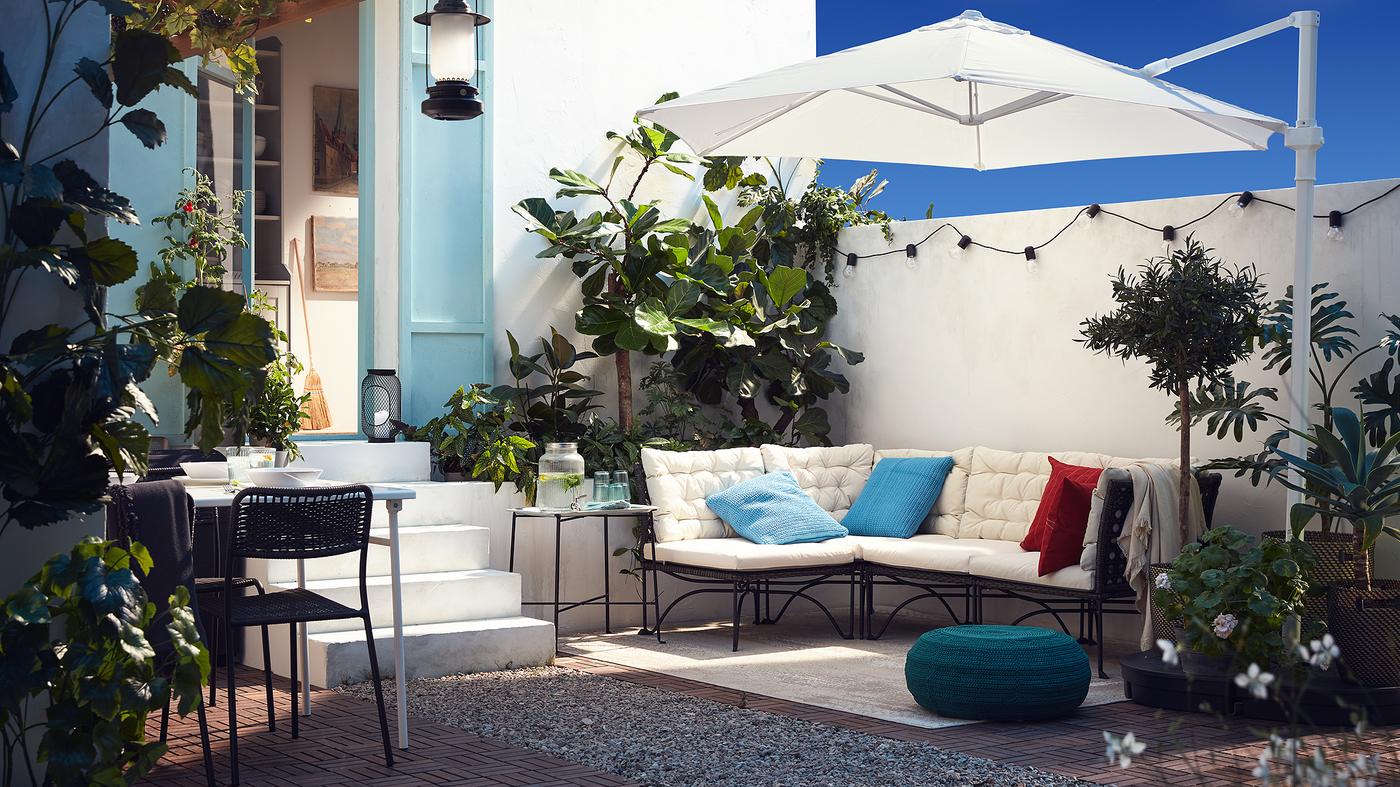 An outdoor patio with a modular sofa with white cushions, a white parasol, a blue-green pouffe and plenty of plants.