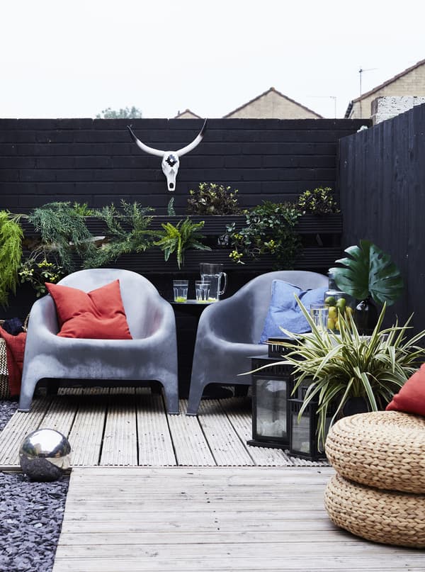 An outdoor terrace with two grey plastic armchairs and rattan pouffes, surrounded with green plants.