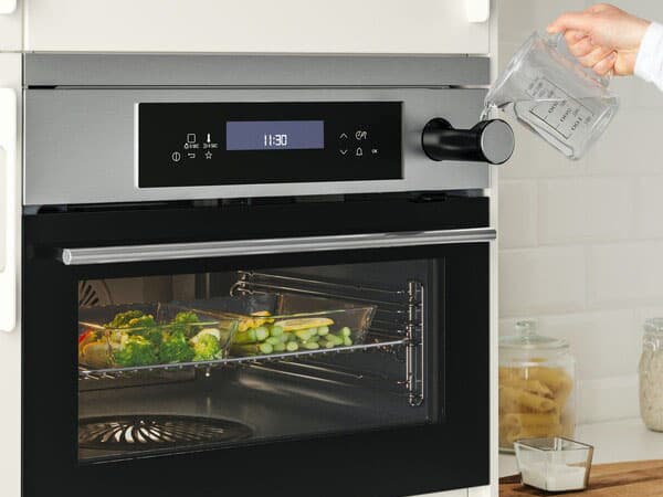 An oven is closed and cooking. A hand holding a jug full of water is pouring it inside a little compartment on the top right of the oven.