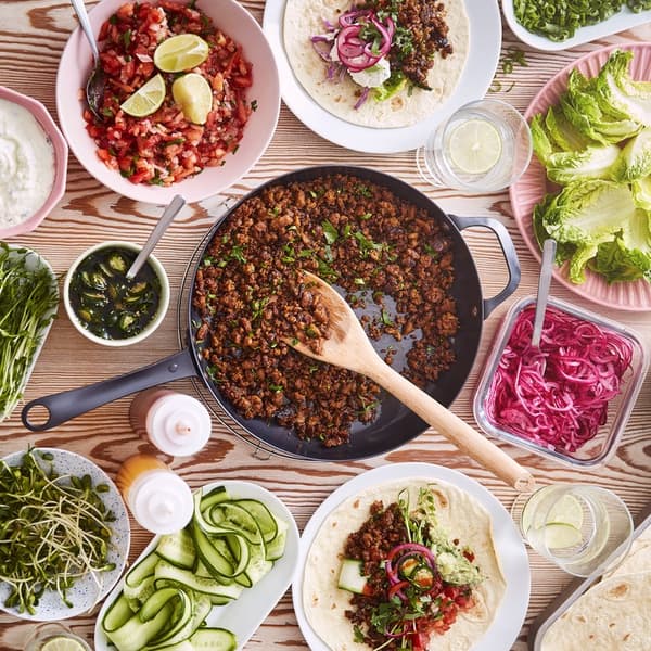 An overhead view of a colourful table spread featuring a central pan of cooked plant-rich mince surrounded by bowls of fresh vegetables, sauces and toppings.				
