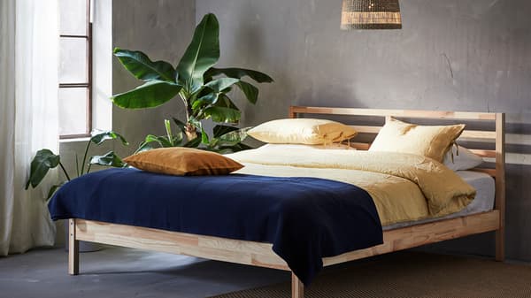 An unmade bed in a bedroom with the windows open, a rug on the floor and lots of green plants.