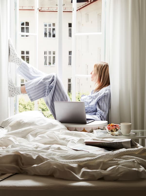 An unmade bed next to an open, sunlit window. On the sill sits a woman, a computer on a BYLLAN laptop support beside her.