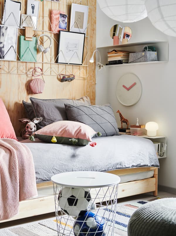 An UTÅKER stackable bed with lots of cushions on, paper art above, a bedside BOTKYRKA shelf, storage and decorations.