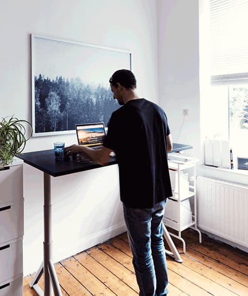 André using a laptop on an adjustable height desk in a room with white walls and a large picture of a forest.