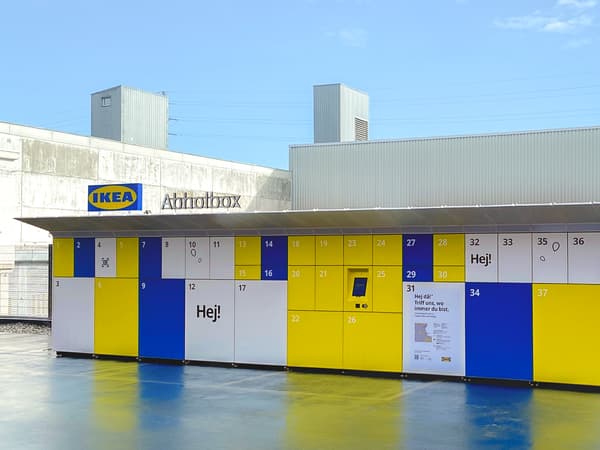 Ansicht einer externen IKEA Abholbox - Einem grossen Container mit diversen nummerierten und verschieden grossen Schliessfächern in den Farben weiss, gelb und blau und einem IKEA Logo auf dem Dach, neben dem der Schriftzug Abholbox angebracht ist. 