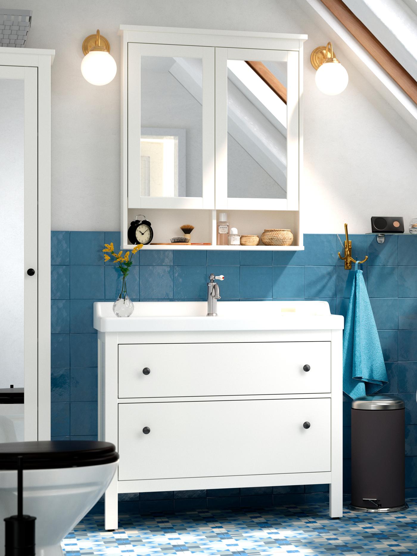  Armarios HEMNES con espejos sobre un lavabo blanco, con azulejos azules detrás y una toalla azul colgando de un gancho.