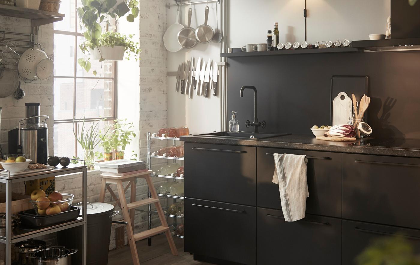 Armoires de cuisine noires avec un évier, une desserte métallique et des ustensiles accroché à des rails à côté d’une fenêtre.