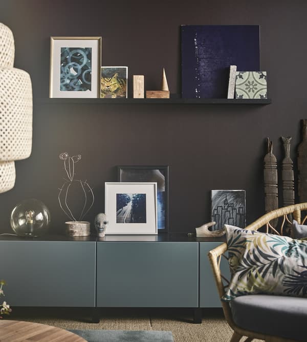 Art arranged on a black picture ledge on a brown wall above a grey-turquoise cabinet in a modern living room.