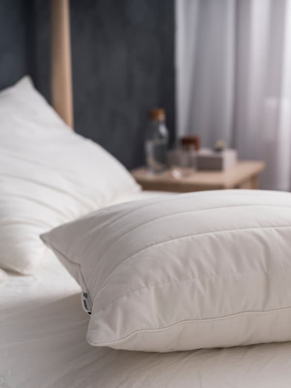 Articles de literie, dont un oreiller ergonomique RUMSMALVA sans taie d’oreiller posé sur un lit en attente de linge de lit propre.