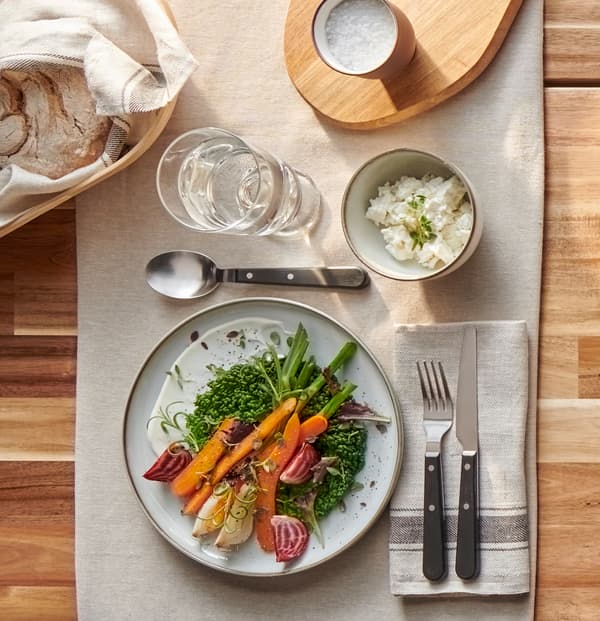 Assiette grise et brune rustique avec légumes colorés, couverts LIVNÄRA robustes et verre d’eau sur une nappe beige.