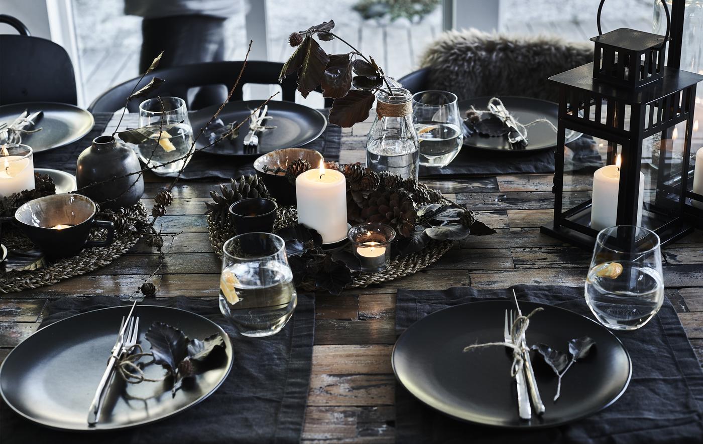 Assiettes noires, couverts en acier, gobelets et centre de table composé d’une bougie bloc allumée et de pommes de pin posées sur un set de table tissé.
