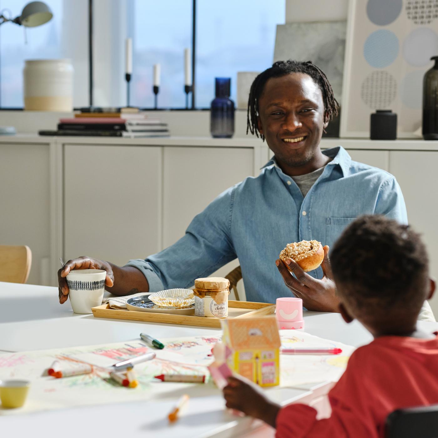 Assis à une table blanche, un adulte tient un mug FRIKOSTIG blanc/à motifs et un petit pain tandis qu’un enfant joue avec des feutres et du papier de la série MÅLA.