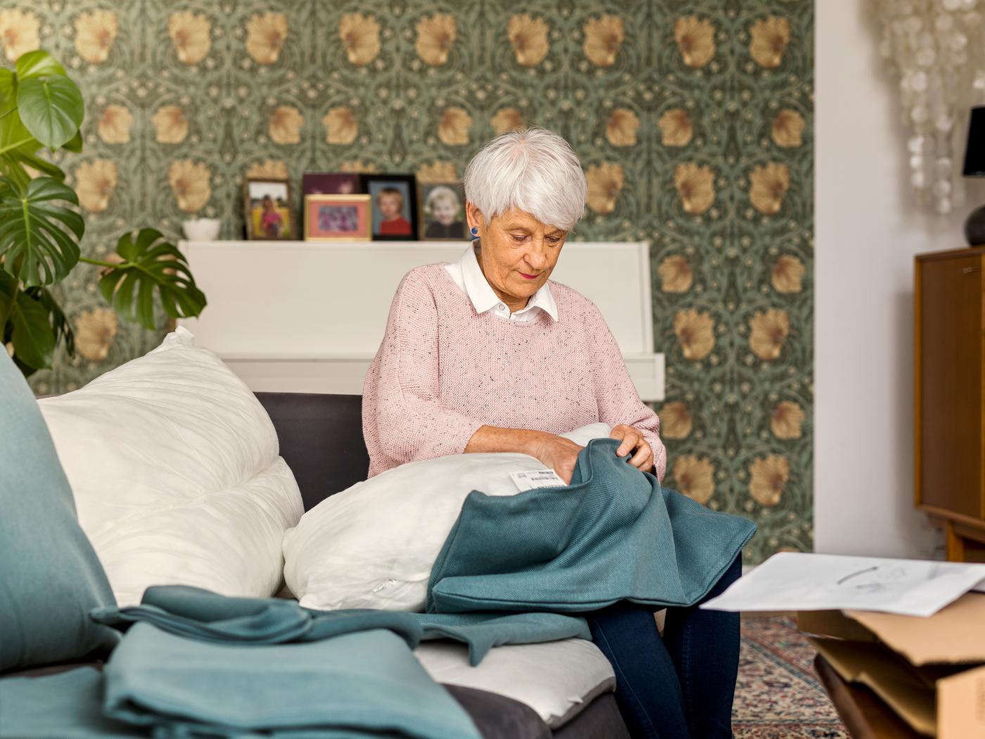 Assise sur un canapé, une femme âgée portant un pull rose clair garnit d’une housse bleue un coussin de canapé.