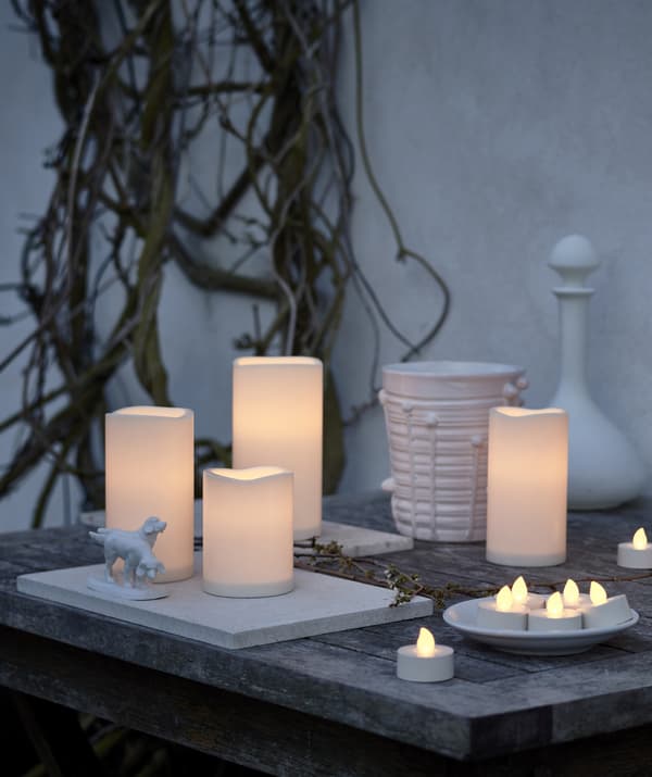 Assortiment de bougies LED rassemblées à l'extérieur sur une table en bois, contre un mur blanc.