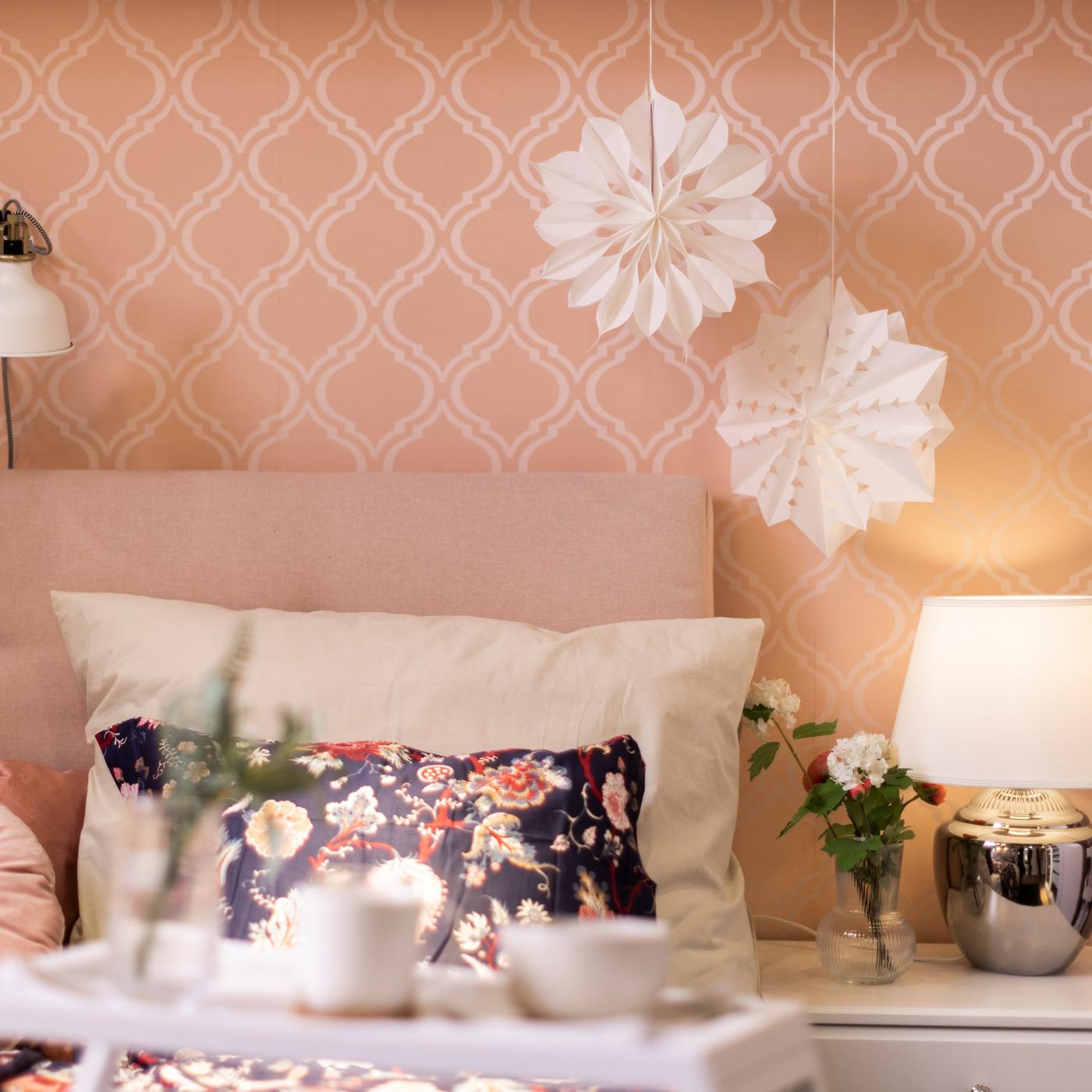 Auf einem Bett liegt ein bunter Polster mit Blumen und einem beige Kissen. Die Wand ist rosa mit einem weißen Muster. Auf der Seite stehen Kästen und eine Tischlampe darauf.