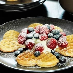 Auf einem schwarzen Teller liegen Waffeln mit Marmelade, Beeren und Schlagobers