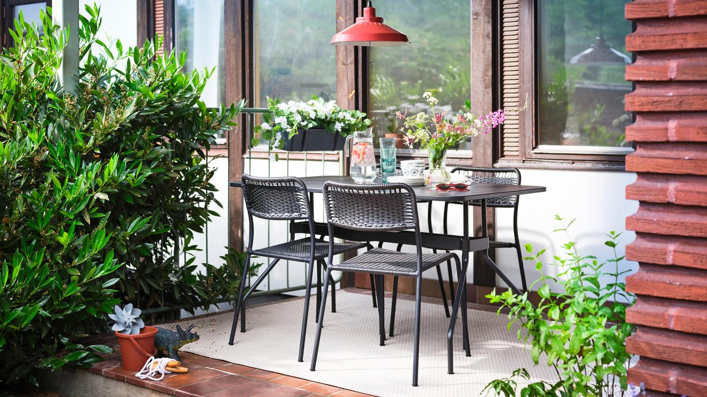 Auf einer Terrasse stehen ein VIHOLMEN Tisch und vier Stühle auf einem cremefarbenen Teppich, u. a. mit einer Vase Blumen, einer Kanne Wasser und Gläsern.