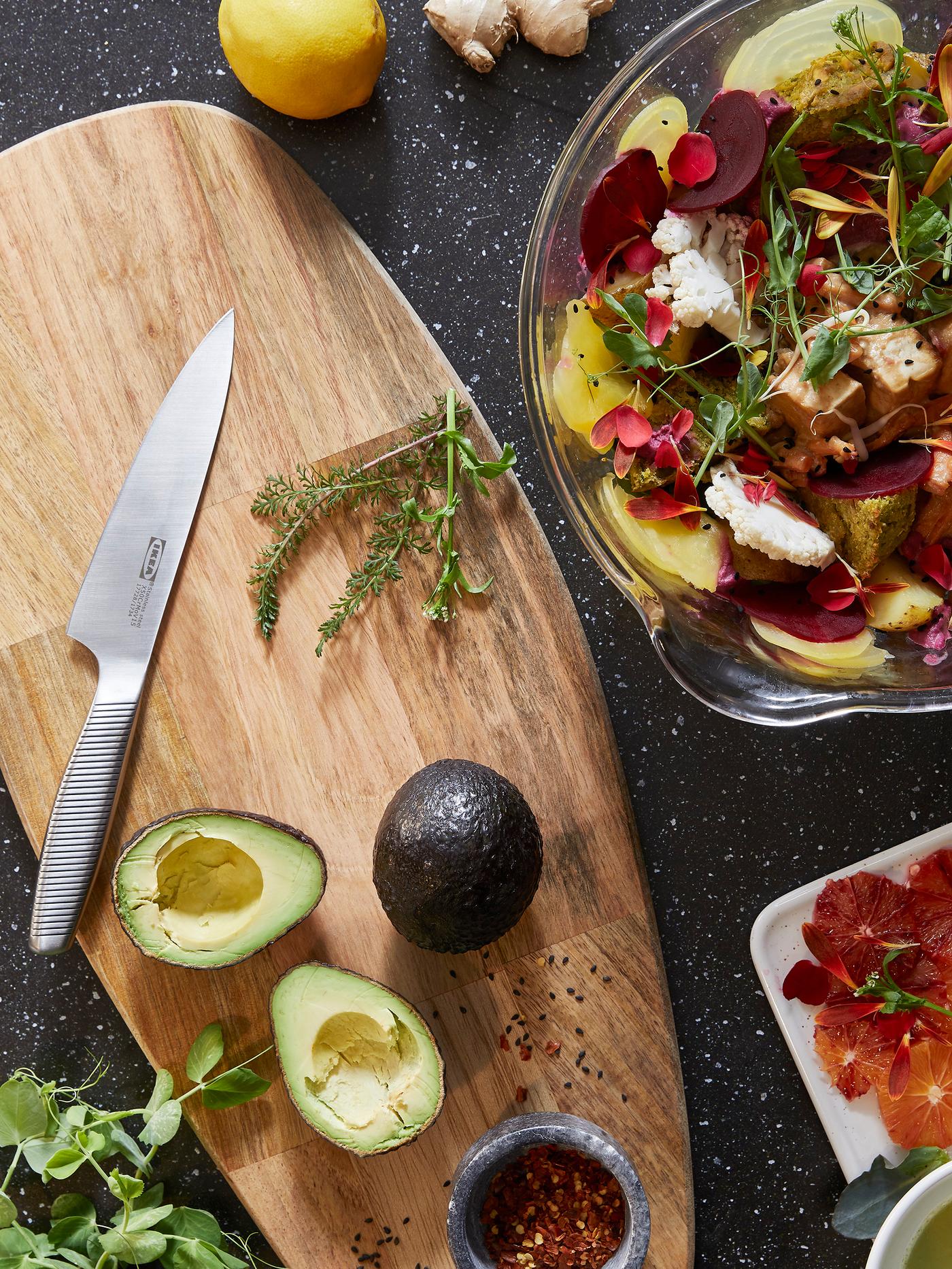 Avokado yang dipotong dan pisau di atas landas cencang kayu bulat FASCINERA IKEA di sebelah mangkuk kaca berisi salad.
