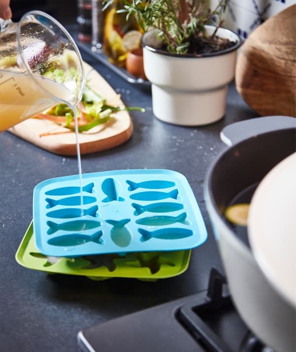 Bacs à glaçons PLASTIS sur un plan de travail de cuisine. Quelqu’un remplit celui du dessus de bouillon de légumes à l’aide d’un gobelet en verre.