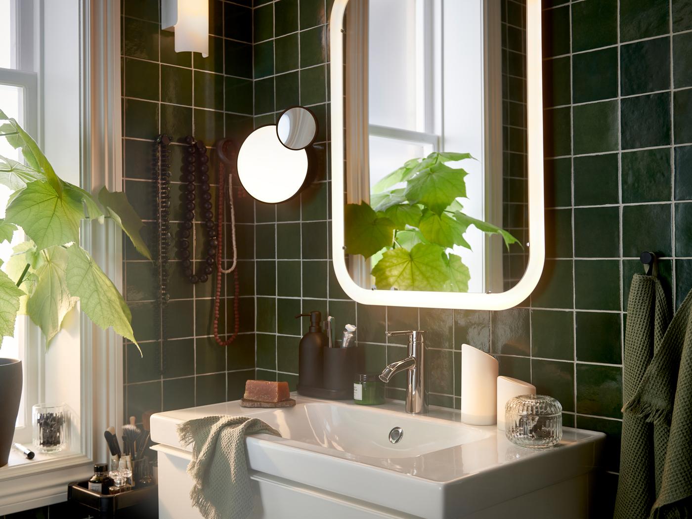 Badkamer met witte spiegel met geïntegreerde verlichting boven een wastafelkast, plus lichtgroene gastendoekjes en een kamerplant.
