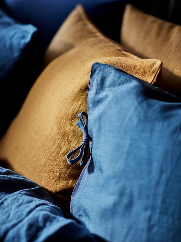 Bahagian sarung bertemu bantal di katil yang sudah dikemas dengan linen katil PUDERVIVA berwarna biru, sepasang kusyen berwarna kuning kunyit gelap terletak antaranya.