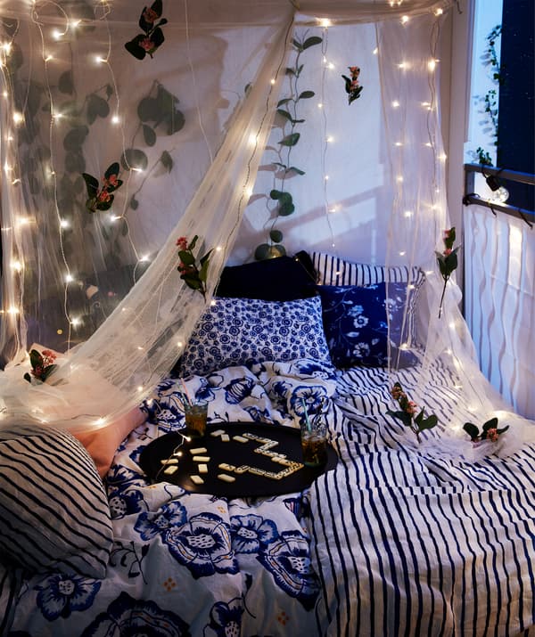 Balcon prêt pour la nuit: lit deux places, boissons servies sur un plateau et dais-moustiquaire garni de guirlandes lumineuses.