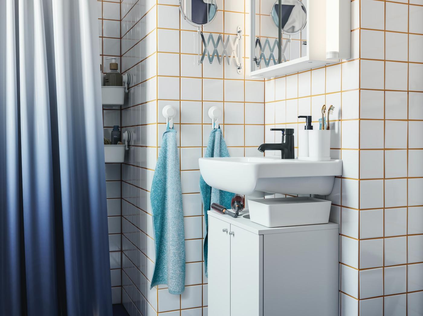 Baño pequeño con los muros cubiertos de cerámicas cuadradas blancas y fragüe amarillo, destacan un pequeño vanitorio separado de un mueble blanco que va abajo, dos toallas de mano celestes y una cortina de ducha en degradé blanco a azul.