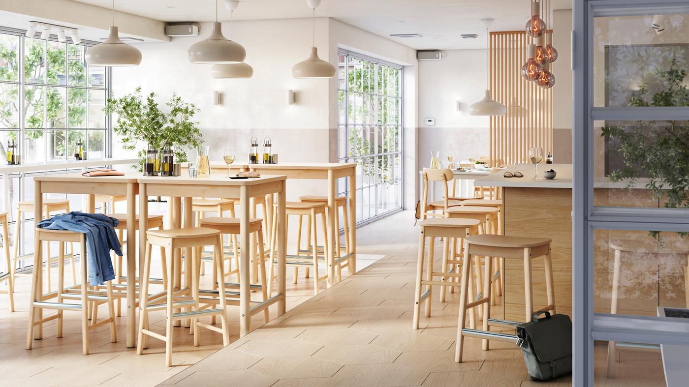 Bar lumineux, aéré et multizones avec tabourets et tables de bar RÖNNINGE près d’une fenêtre, et tabourets de bar devant un bar au centre.